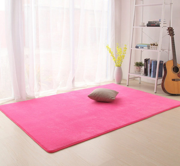 Ce tapis est rose foncé et rectangulaire. Il est placé sous une fenêtre dans une chambre. Un coussin est posé dessus.