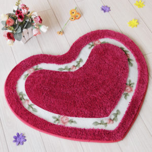 Paillasson rouge en forme de cœur avec ligne blanche et fleurs vertes et rouge. Il est posé sur un parquet clair avec ces fleurs colorés.