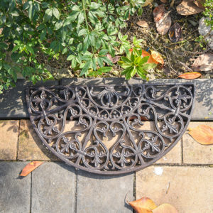 Ce paillasson en fonte est en forme de demi-cercle. Il est placé sur un sol en briques et devant une plante.