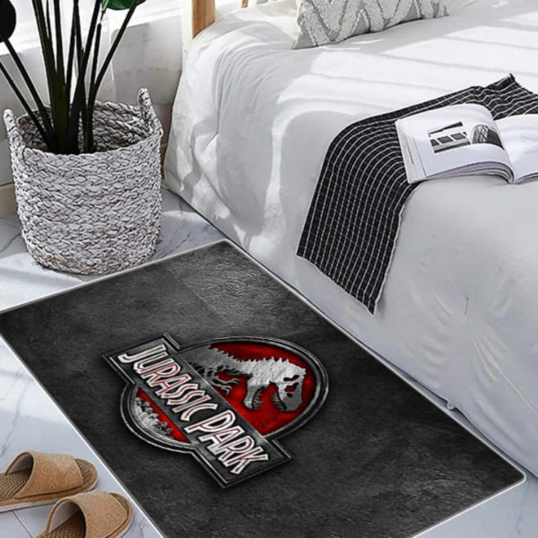 Paillasson Jurassic Park gris avec logo rouge. Il est dan une pièce avec un lit, des chaussons et une plante.