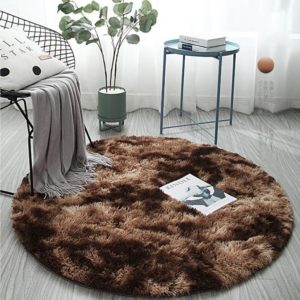 Ce tapis rond est marron. Il est placé sur un sol gris. Il y aune chaise, une plante et une petite table ronde.