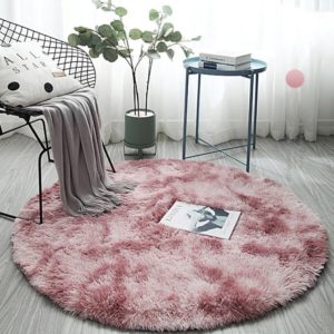 Ce tapis rond est rose. Il est placé sur un sol gris. Il y aune chaise, une plante et une petite table ronde.