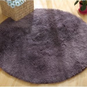 Ce tapis rond est violet . Il est disposé sur du parquet. Un casier en osier avec des fleurs est placé sur le coin supérieur gauche.