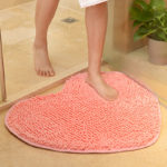 Un paillasson rose clair en forme de coeur est posé au sol dans une salle de bain. Une femme en serviette sort de la douche et pose son pied sur le paillasson. Une plante est posée sur sol à côté de la douche.