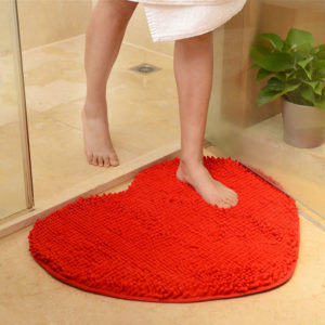Un paillasson rouge en forme de coeur est posé au sol dans une salle de bain. Une femme en serviette sort de la douche et pose son pied sur le paillasson. Une plante est posée sur sol à côté de la douche.