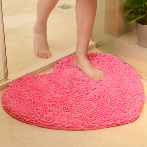 Un paillasson rose en forme de coeur est posé au sol dans une salle de bain. Une femme en serviette sort de la douche et pose son pied sur le paillasson. Une plante est posée sur sol à côté de la douche.
