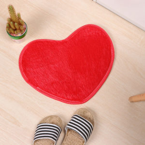 Ce tapis rouge est en forme de coeur . Il est placé sur du parquet avec une plante verte et des claquettes.