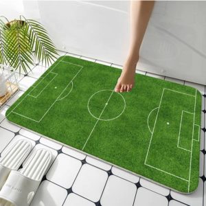 paillasson vert imprimé comme un terrain de foot. Le tapis est posé sur un carrelage avec des chaussons à coté et une plante. Quelqu'un sort de son bain sur ce tapis