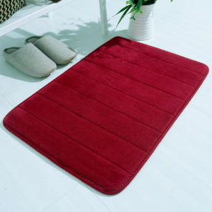 Un tapis rouge antidérapant et agréable au toucher posé sur un sol blanc avec des chaussons blancs à côté et une plante.